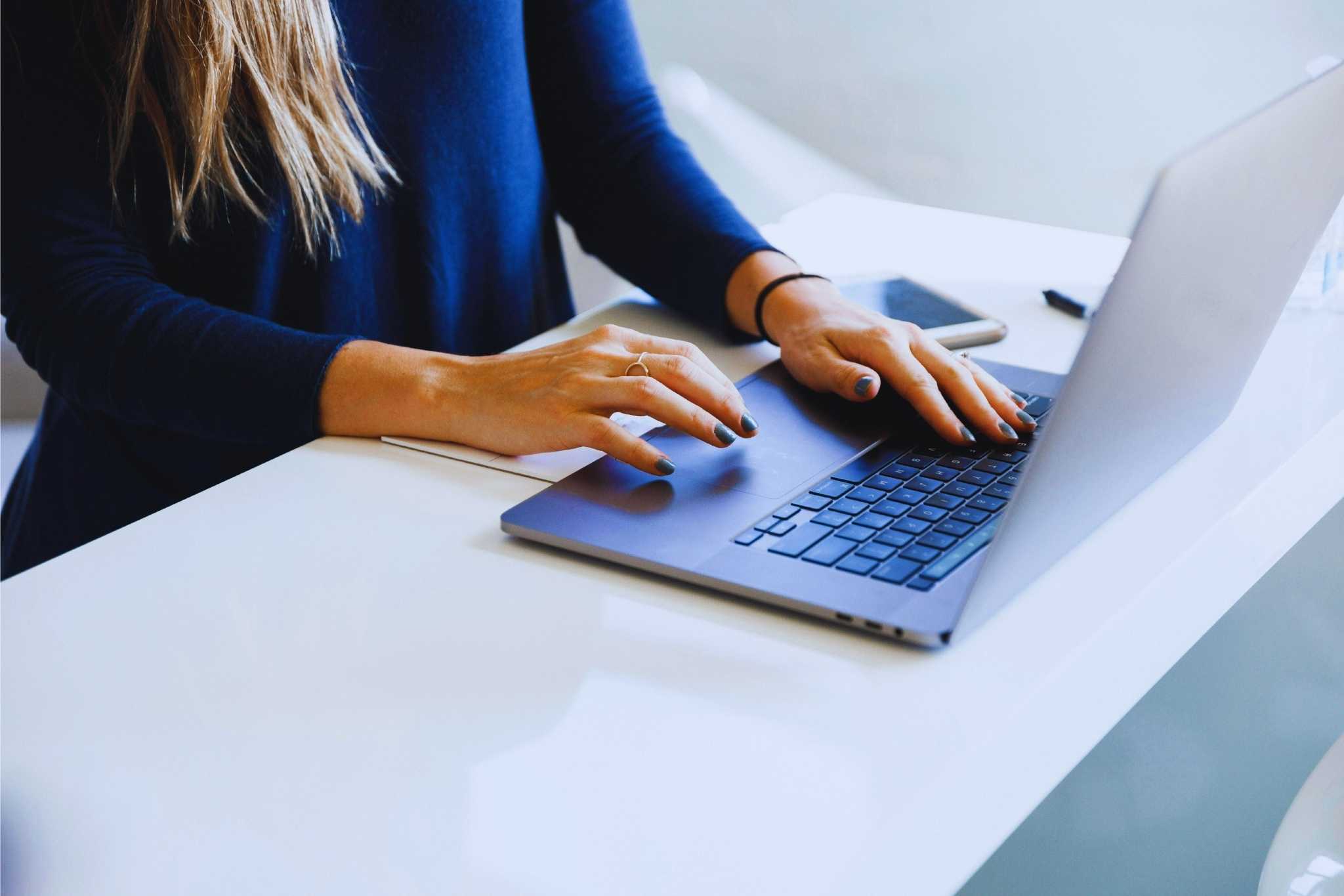 Person mit langem Haar und blauem Oberteil arbeitet am Laptop in einem modernen, aufgeräumten Büro mit Smartphone und Notizbuch.