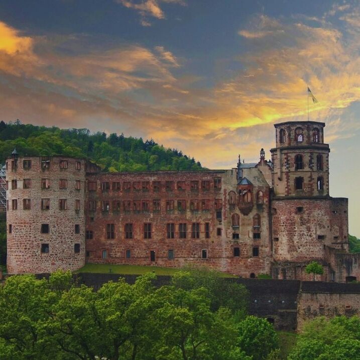 Heidelberger Schloss bei Sonnenuntergang, umgeben von grüner Hanglandschaft – historisches Wahrzeichen mit rötlicher Sandsteinfassade und markanten Türmen.