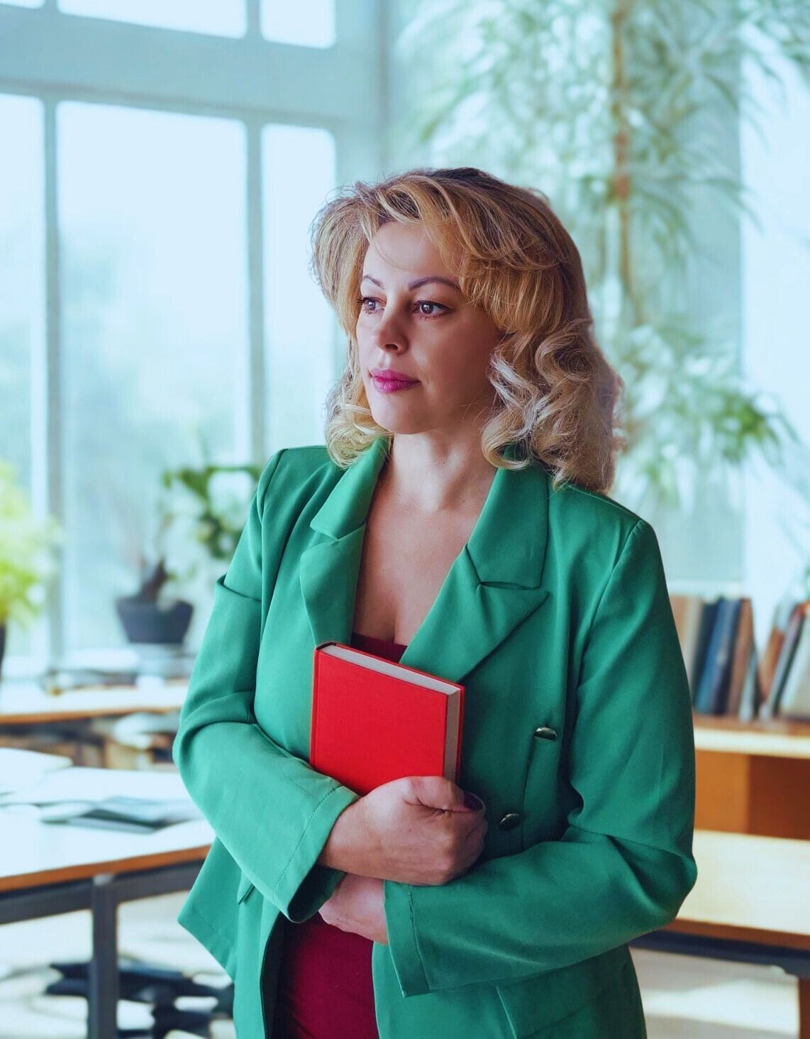 Über uns Ivana Cvetkovic, Geschäftführerin in modernem Büro mit grünen Pflanzen und großen Fenstern, selbstbewusst mit einem Buch in den Armen stehend.