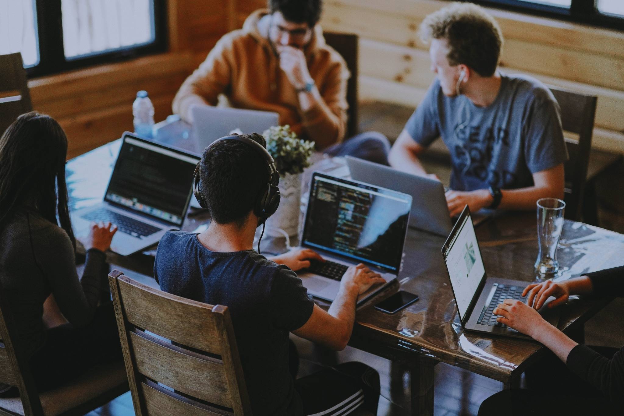 Gruppe junger Menschen arbeitet gemeinsam an Laptops an einem Holztisch in einem hellen Raum mit großen Fenstern.