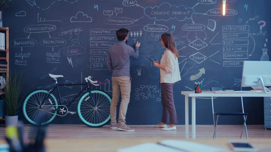 Gebäudereinigung Zwei Personen stehen vor einer großen beschrifteten Tafelwand mit Diagrammen und Notizen in einem modernen Büro.