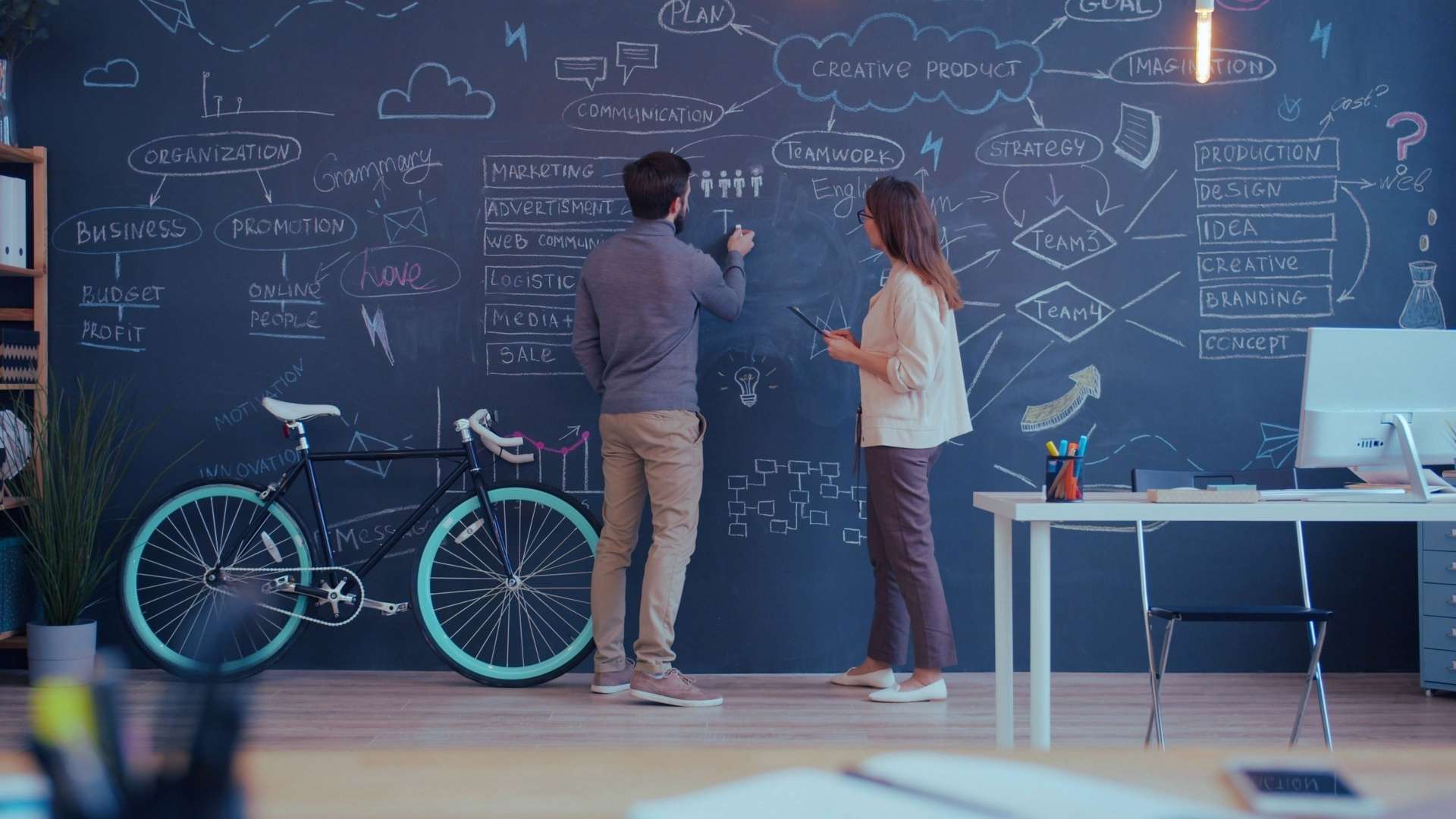 Zwei Personen stehen vor einer großen beschrifteten Tafelwand mit Diagrammen und Notizen in einem modernen Büro.